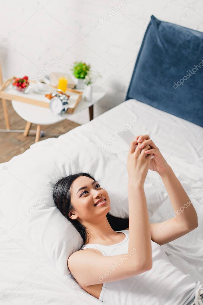 High angle view of cheerful asian woman using smartphone while lying on bed 