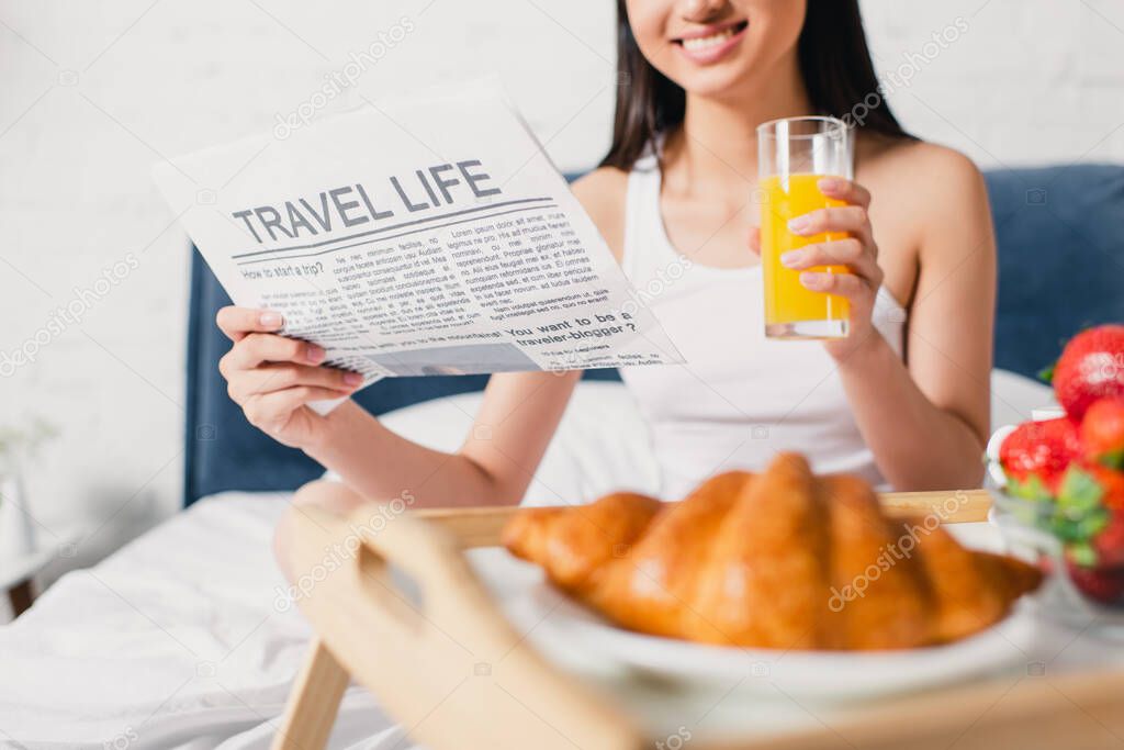 Cropped view of smiling woman holding newspaper with travel life lettering and glass of orange juice on bed at morning 