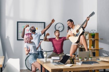 Ofiste şarkı söyleyip dans eden çok kültürlü iş arkadaşlarının yanında akustik gitar çalan iş kadınının seçici odağı 