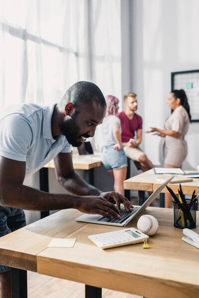 Селективный фокус африканского американского бизнесмена, использующего ноутбук во время работы рядом с многонациональными коллегами в офисе 