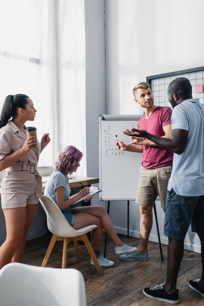Селективный фокус бизнесмена, указывающего на доску возле мультикультурных коллег с кофе, чтобы пойти и буфер обмена в офисе 