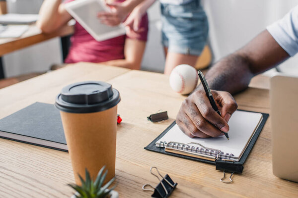 Селективный фокус африканского американского бизнесмена, пишущего на планшете возле ноутбука, кофе, чтобы пойти и канцелярские принадлежности на столе