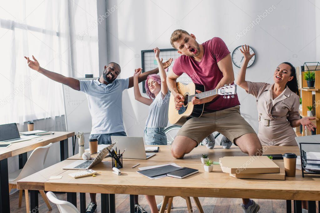 Selective focus of multicultural business people with acoustic guitar dancing in office 
