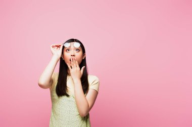 shocked asian girl looking up, covering mouth and touching glasses isolated on pink  clipart