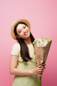 mladá asijská žena v slamáku s úsměvem a drží květiny izolované na růžové 