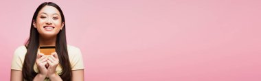 Pembe üzerinde kredi kartı tutan neşeli ve genç Asyalı kadının yatay görüntüsü. 