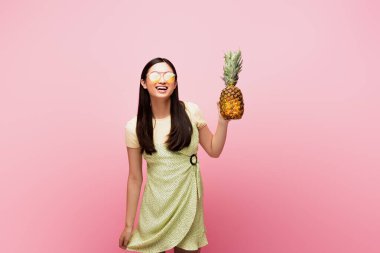 happy asian girl in sunglasses holding ripe pineapple isolated on pink  clipart