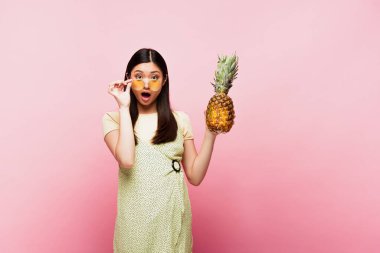 Şok olmuş güneş gözlüklü Asyalı kız pembe üzerinde olgun ananas tutuyor. 