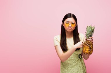 Güneş gözlüklü üzgün Asyalı kadın pembe üzerinde taze ananas tutuyor. 
