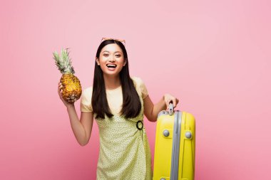 Mutlu Asyalı kız pembe üzerine izole edilmiş bavul ve taze ananas taşıyor. 