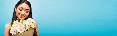 Mutlu Asyalı kızın panoramik mahsulü gülümsüyor ve mavi üzerinde çiçekler tutuyor.