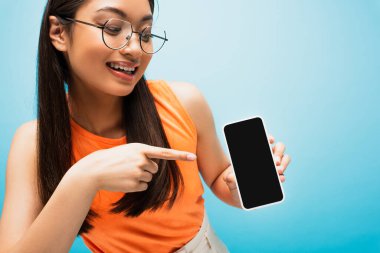 Gözlüklü neşeli Asyalı kız mavi ekranlı akıllı telefonu işaret ediyor.