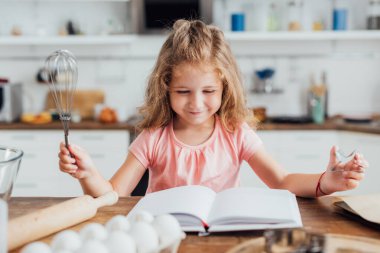 selective focus of child holding whisk while reading cookbook near chicken eggs and rolling pin clipart