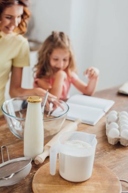 Anne ve kızının yumurta, süt, un, cam kase ve yemek pişirme aletlerinin yanında yemek kitabı okuduğu seçici odak noktası