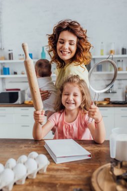 selective focus of girl holding rolling pin and sieve near mother with infant son clipart