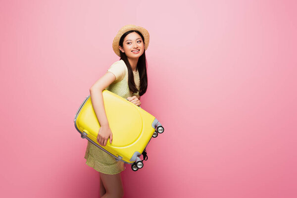cheerful asian girl in straw hat holding luggage on pink 