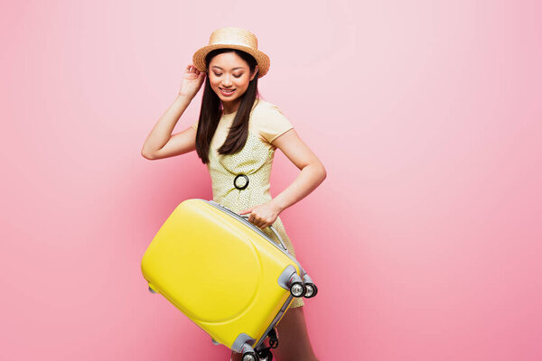 positive asian girl in straw hat looking at yellow luggage on pink 