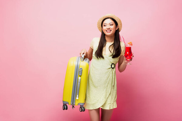 cheerful asian girl in straw hat holding cocktail and yellow baggage on pink