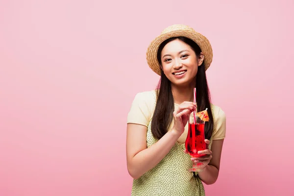 笑容满面的亚洲女人头戴草帽 手里拿着酒杯 鸡尾酒与粉色隔离 — 图库照片