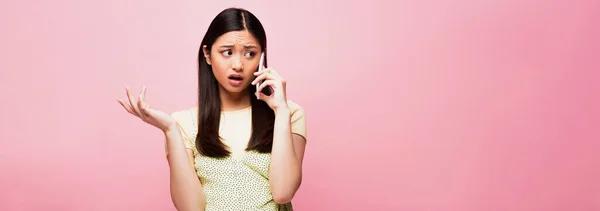 口を開いてジェスチャーし ピンクに隔離されたスマートフォンで話している不満アジアの女性のパノラマショット — ストック写真