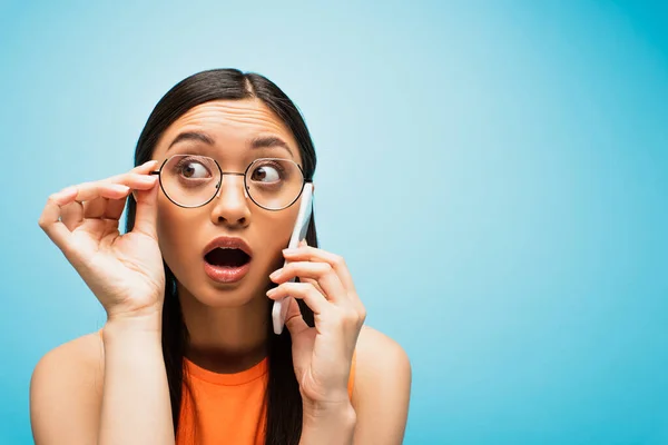 Chocado Asiático Menina Tocando Óculos Enquanto Falando Smartphone Azul — Fotografia de Stock