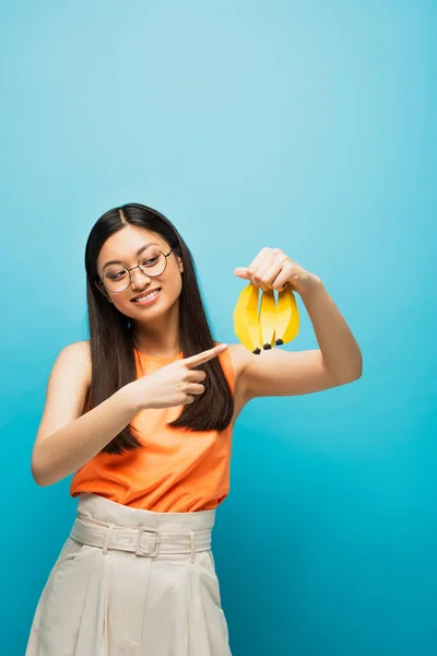 Cheerful Asian Girl Glasses Pointing Finger Ripe Bananas Isolated Blue — Stock Photo, Image