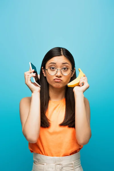 Confused Asian Girl Glasses Holding Smartphone Banana Isolated Blue — Stock Photo, Image
