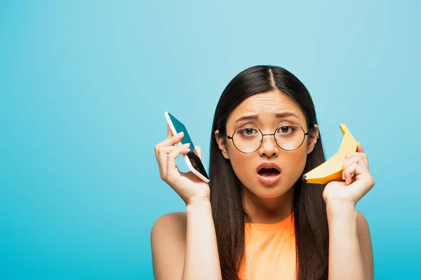 Conmocionado Chica Asiática Gafas Celebración Teléfono Inteligente Plátano Aislado Azul — Foto de Stock