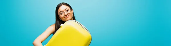 Poziomy Obraz Wesołej Azjatki Okularach Przytulających Żółty Bagaż Niebiesko — Zdjęcie stockowe