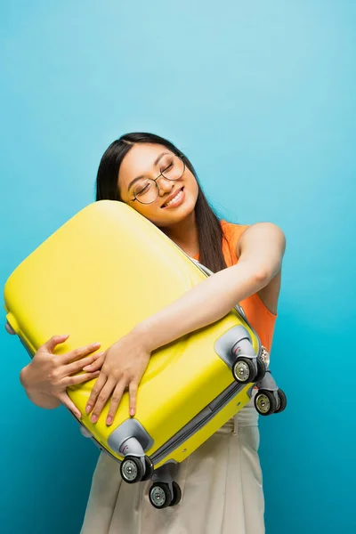 Cheerful Asian Woman Glasses Hugging Yellow Luggage Blue — Stock Photo, Image