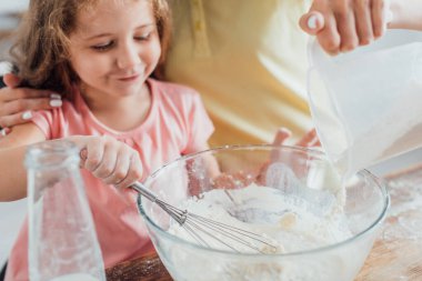 Annenin elinde sürahi tutması ve kızının hamur yoğurması, seçici bir odak noktası.
