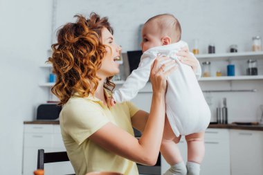 Genç, kıvırcık anne, mutfakta bebeği tutuyor.