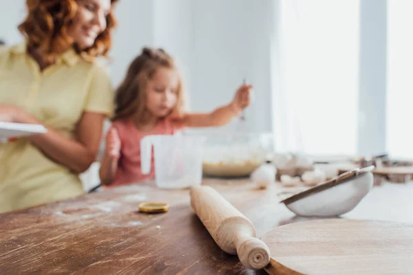 selective focus of chopping board, rolling pin and sieve near mother and daughter cooking in kitchen