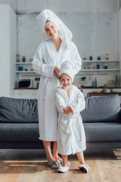 молодая мать и дочь в белых махровых халатах и полотенцах на головах смотрят в камеру, стоя рядом с диваном