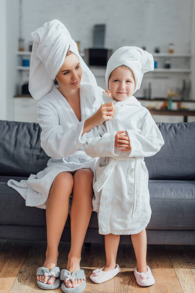 молодая мама сидит на диване и трогает дочь в халате и полотенце на голове