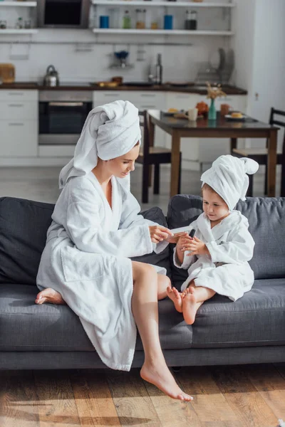 young woman making manicure with nail file to daughter while sitting together in white bathrobes and towels on heads