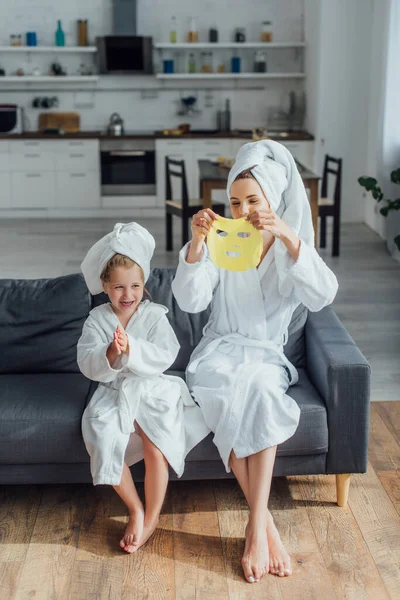 Young Woman Holding Face Mask While Sitting Sofa Daughter Bathrobes — Stock Photo, Image