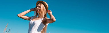 low angle view of young blonde woman touching straw hat while looking away against blue sky, horizontal image clipart