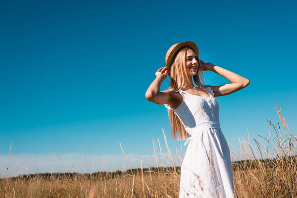 selective focus of woman in white dress touching straw hat while looking away in grassy meadow against blue sky