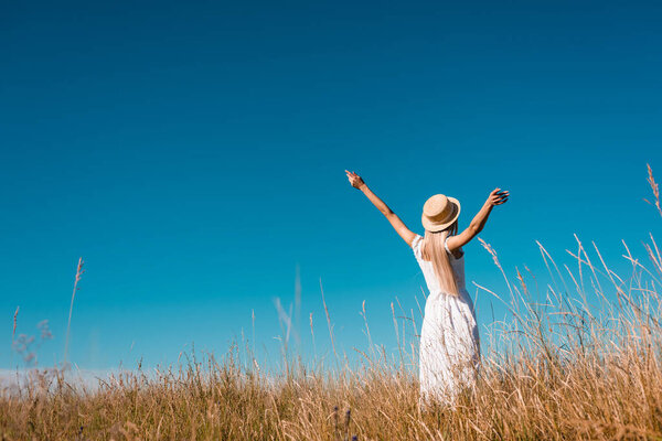 Вид сзади женщины в белом платье и соломенной шляпе, стоящей с протянутыми руками на фоне голубого неба