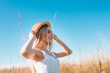 selective focus of stylish blonde woman touching straw hat while looking away against blue sky  clipart