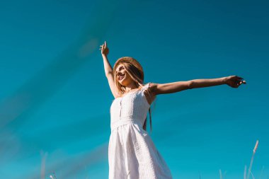 Düşük açılı beyaz elbiseli kadın ve hasır şapkalı kadın mavi gökyüzüne uzanmış uzanmış ellerle bakarken başka tarafa bakıyor.