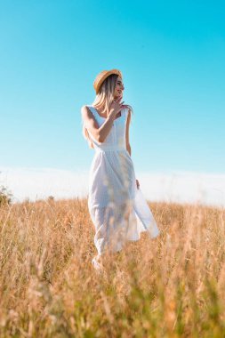 Beyaz elbiseli ve hasır şapkalı şık bir kadının seçici odağı. Çeneye dokunuyor ve mavi gökyüzüne bakıyor.