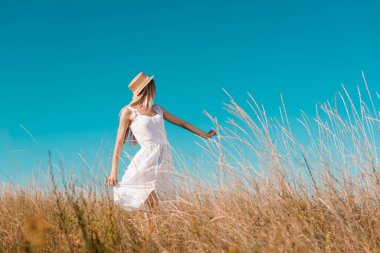 selective focus of blonde woman in white dress and straw hat showing follow me gesture on grassy meadow clipart