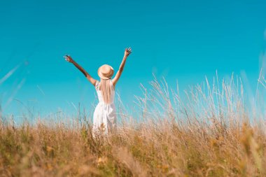 back view of woman in straw hat and white dress standing with raised hands on grassy meadow, selective focus clipart