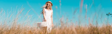 selective focus of blonde woman in white dress and straw hat standing on wind in field, horizontal concept clipart