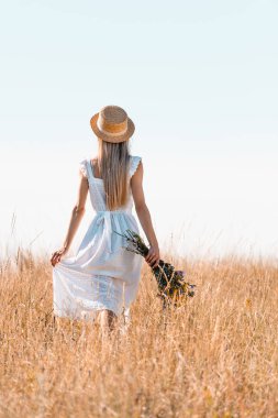 Hasır şapkalı bir kadının beyaz elbiseye dokunuşunun arka planında bir buket kır çiçeğiyle çimenli çayırda yürürken