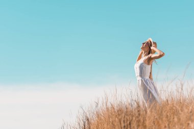 selective focus of young woman in white dress touching straw hat while standing on grassy hill against blue sky clipart