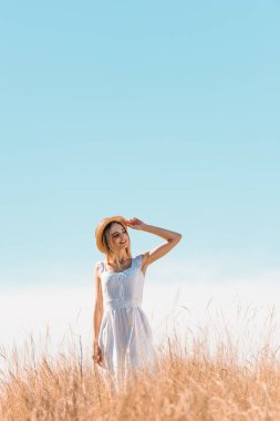 Beyaz elbiseli genç kadın çimenli tepede duruyor, hasır şapkaya dokunuyor ve mavi gökyüzüne bakıyor.