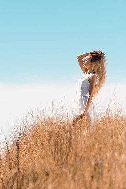 Seçici odak noktası beyaz elbiseli, saçlarına dokunan ve saman şapkalı genç bir kadın çimen tarlasında kapalı gözlerle dikilirken